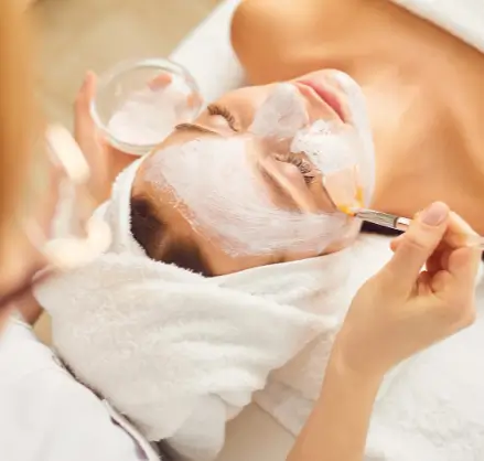 A client at Serenity Head Spa receives a facial treatment. A therapist uses a brush to apply a white mask to the client's face, who has their eyes closed and is wrapped in white towels.