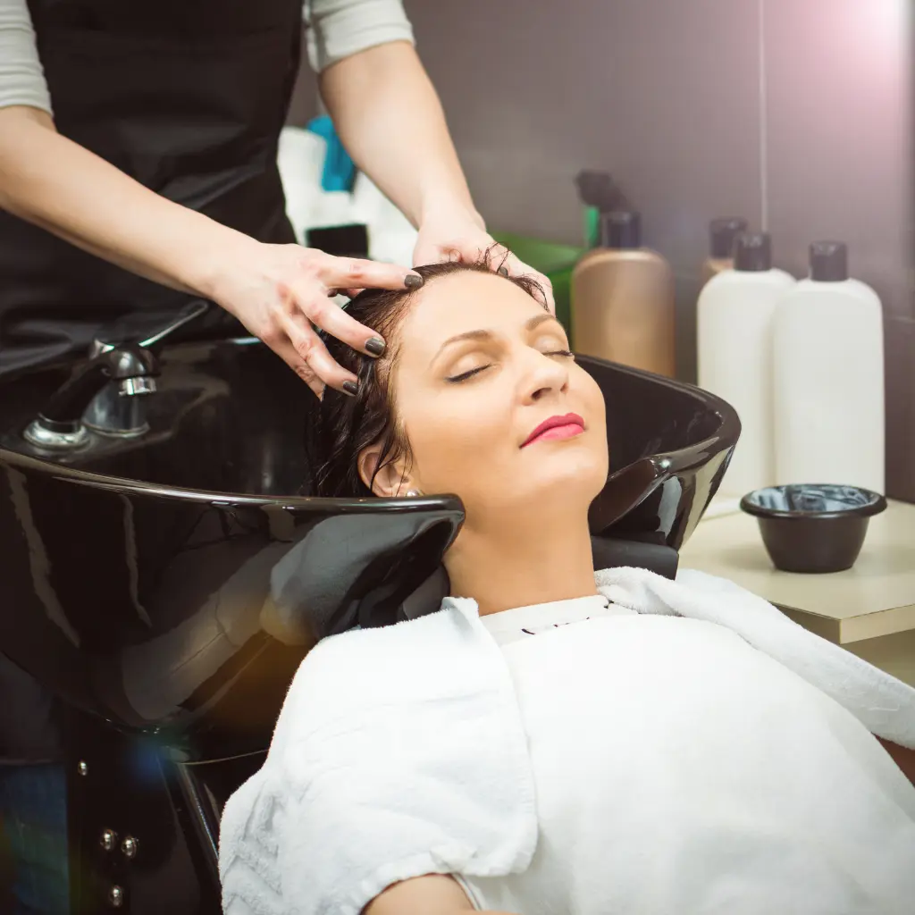 A Woman With Her Eyes Closed, Leaning Back In A Salon Sink, Getting Her Hair Washed By A Hairdresser.