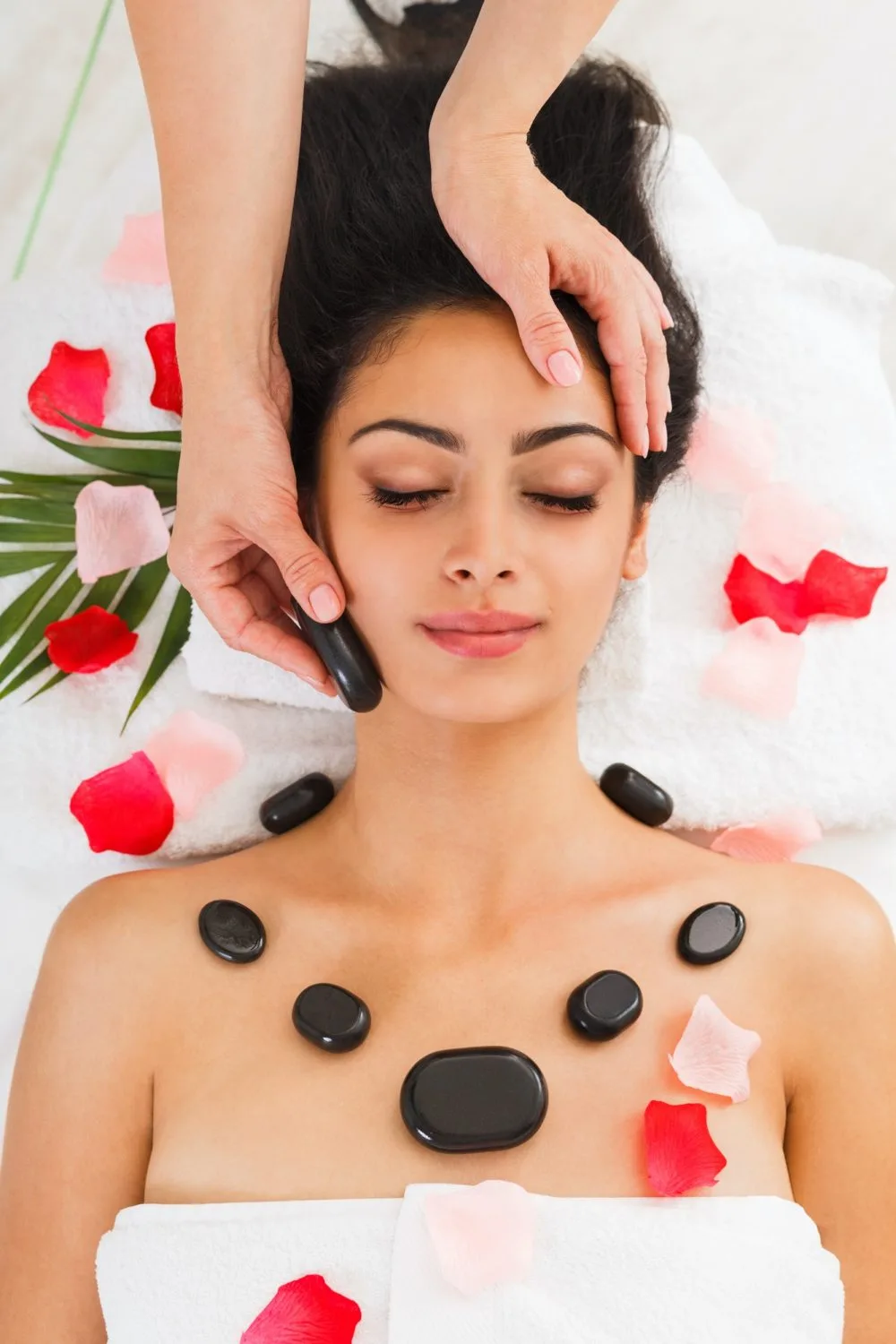 A woman with a bare upper body lying on a white surface, receiving a hot stone massage with black stones and flower petals.