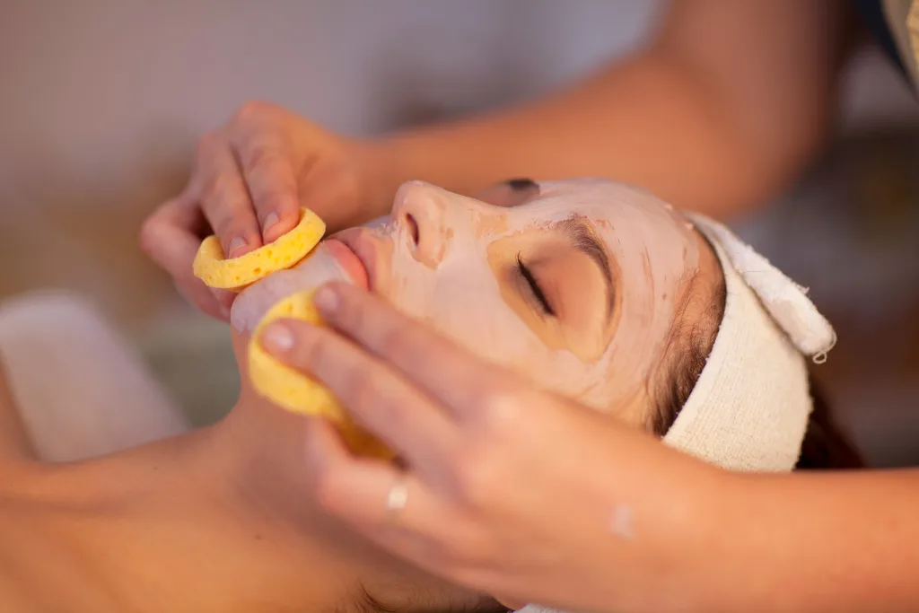 A client with a facial mask on is receiving a treatment at Serenity Head Spa. Two hands are gently using yellow sponges to work on the client's face.