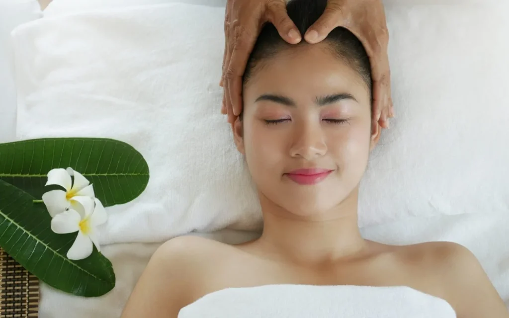 A serene scene at Serenity Head Spa. A relaxed client with closed eyes receives a gentle head massage. White frangipani flowers and green leaves adorn the treatment area.