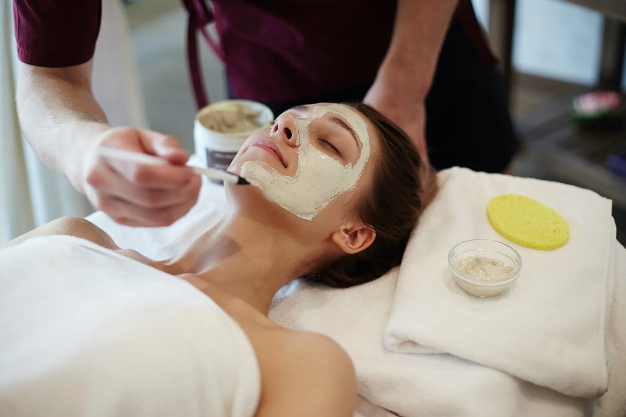 A client with closed eyes receives a facial mask application at Serenity Head Spa. A therapist uses a brush to apply a light-colored mask to the client's face. A bowl of product and a yellow sponge rest on a nearby towel.