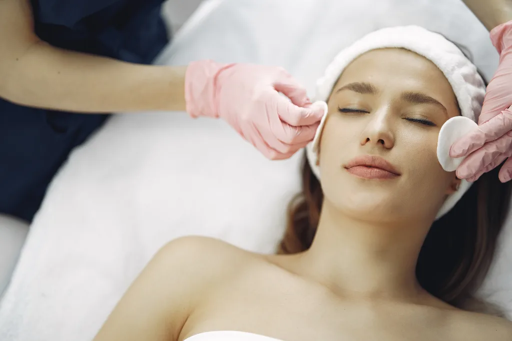 A client with closed eyes and a white headband receives a facial cleansing at Serenity Head Spa. Aestheticians wearing pink gloves gently use cotton pads on the client's face.