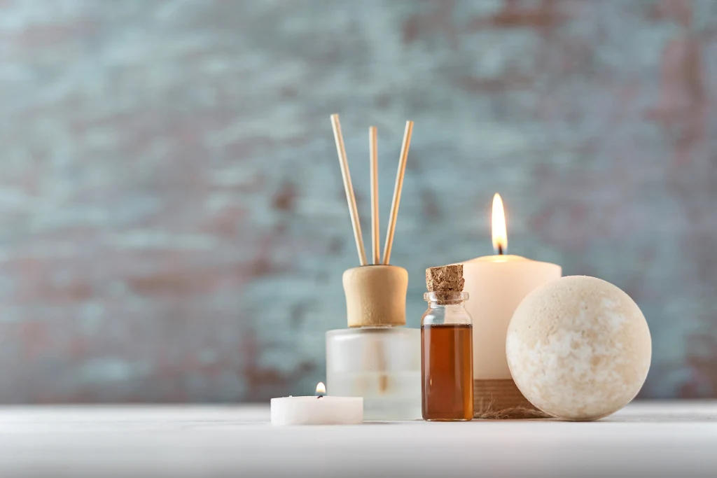 A collection of spa products including a reed diffuser, a small vial of essential oil, a lit candle, and a bath bomb against a rustic blue background.