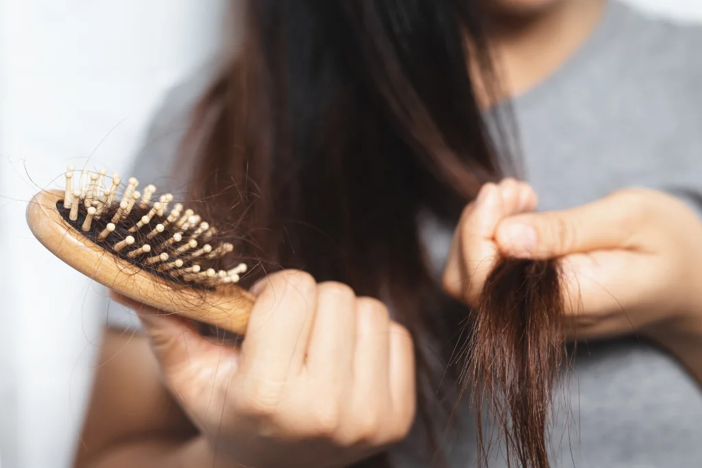 A person holding a wooden hairbrush filled with loose hair strands while touching their long, dark hair.