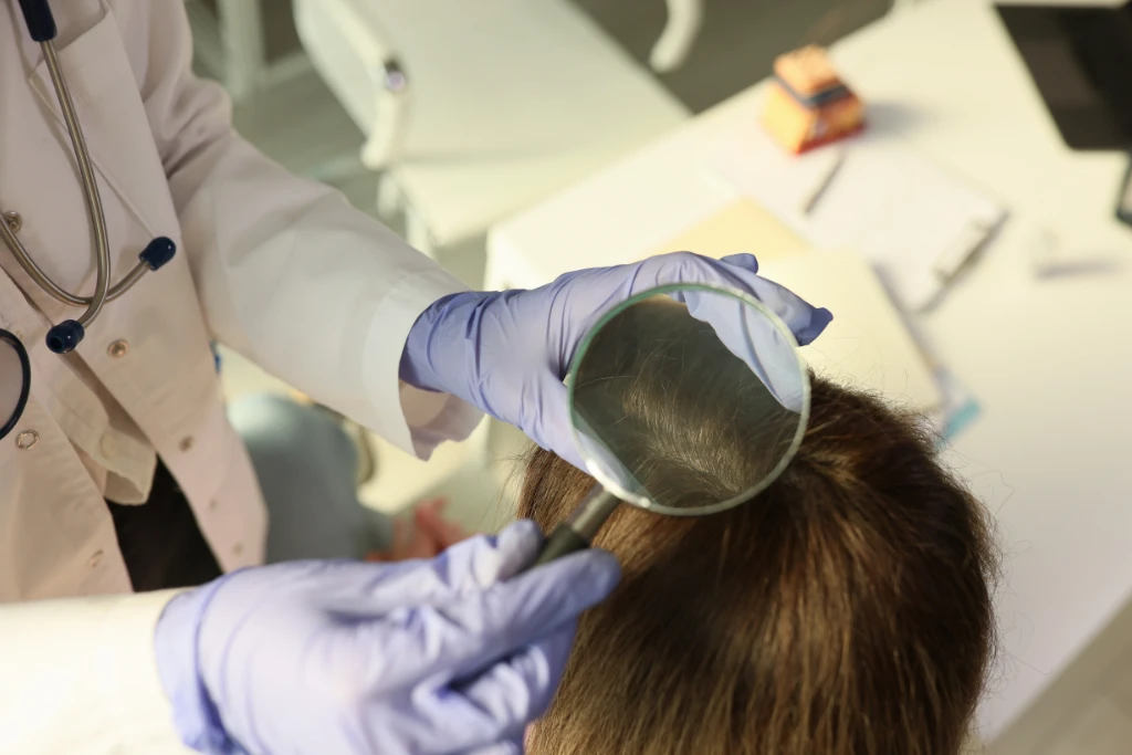 A close-up, top-down view of a specialist in a white lab coat and purple gloves using a magnifying glass to inspect a client's scalp and hair density.