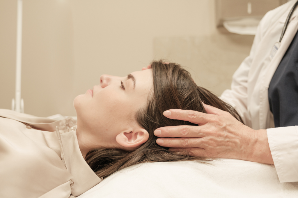 A woman lying down with her eyes closed while a healthcare professional in a white lab coat performs a therapeutic scalp massage.