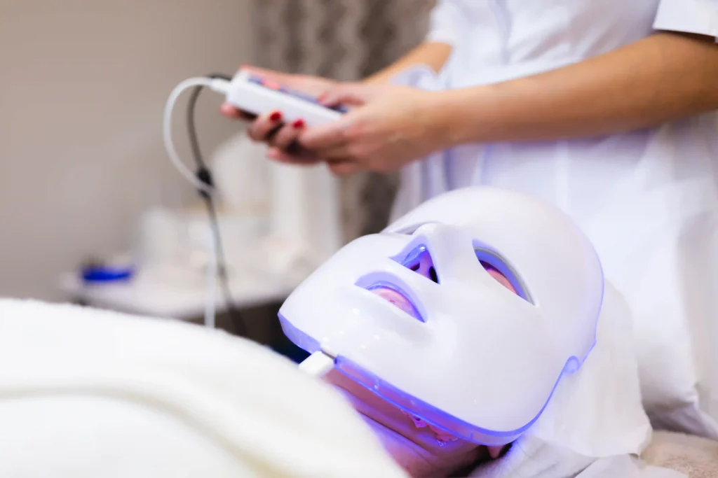 A close-up of a client wearing a white LED light therapy facial mask with blue light while a specialist operates the control device.