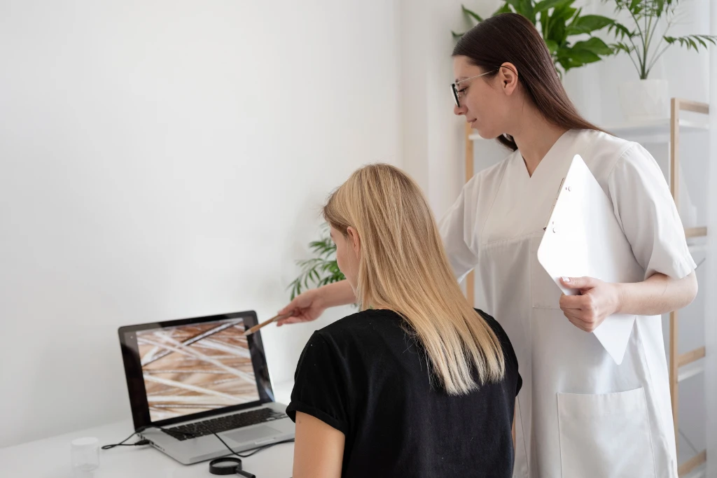 A trichologist or hair specialist in a white uniform pointing at a laptop screen to show a blonde client a microscopic view of their hair follicles and scalp health.