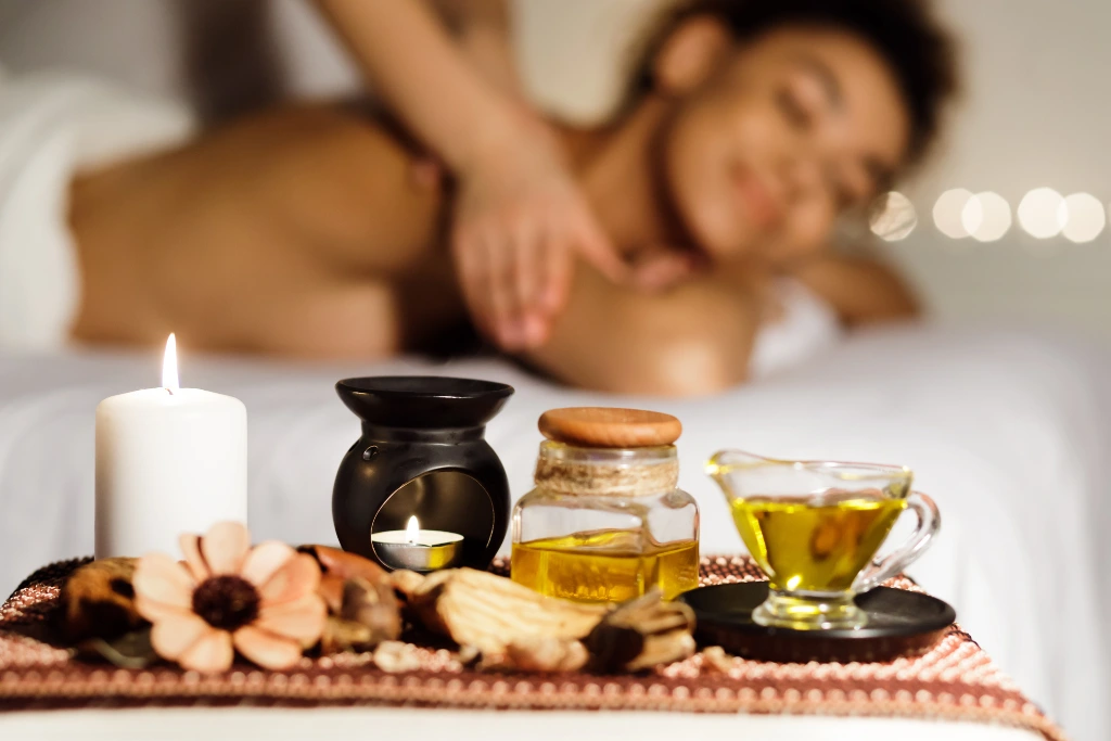 A Tray Of Massage Essential Oils, An Oil Burner, And Candles In The Foreground With A Woman Receiving A Relaxing Aromatherapy Massage Blurred In The Background.