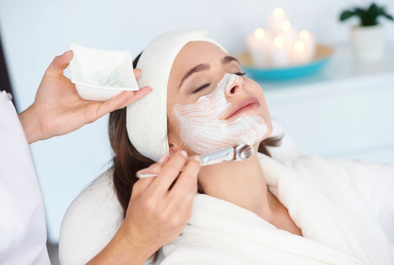 woman relaxing with her eyes closed while an aesthetician applies a white hydrating facial mask using a professional brush.