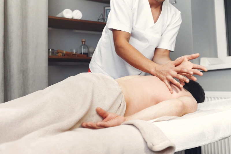 A Male Massage Therapist Or Physiotherapist In White Scrubs Performs A Deep Tissue Back Massage On A Male Client Lying On A Treatment Table.
