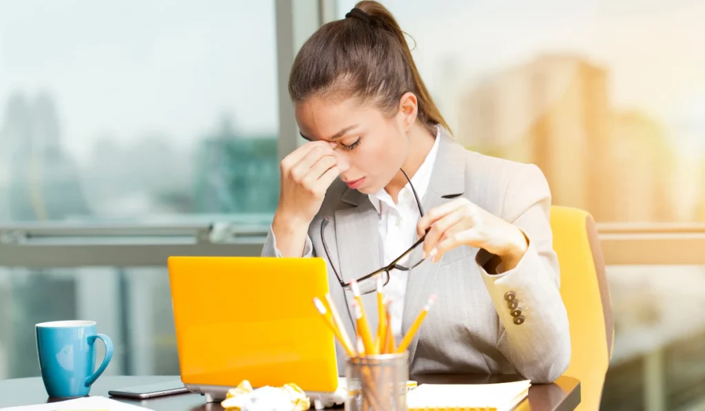 Massage For Stress Relief: A Simple Guide To Calming Your Mind And Body 1 A Professional Woman Rubbing Her Eyes And Taking Off Her Glasses While Working On A Laptop At A Bright Office Desk, Experiencing Fatigue Or A Migraine.
