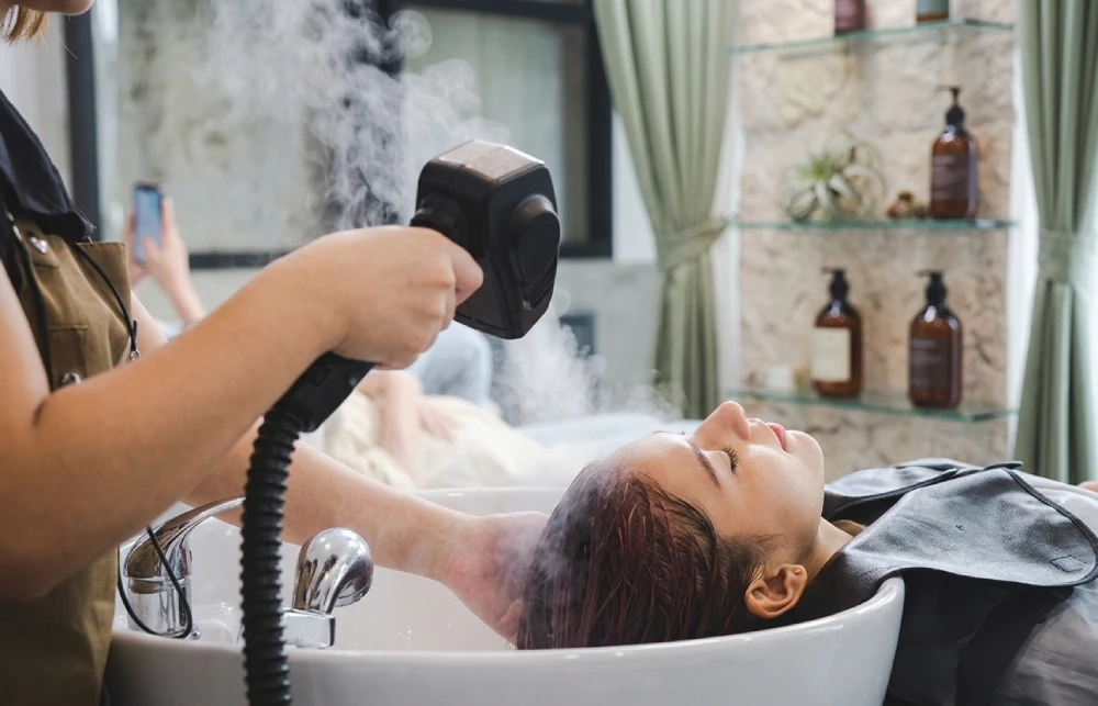 A Woman Lying Down At A Head Spa Sink Receiving A Hair Steaming Treatment From A Therapist Holding A Mist Wand.